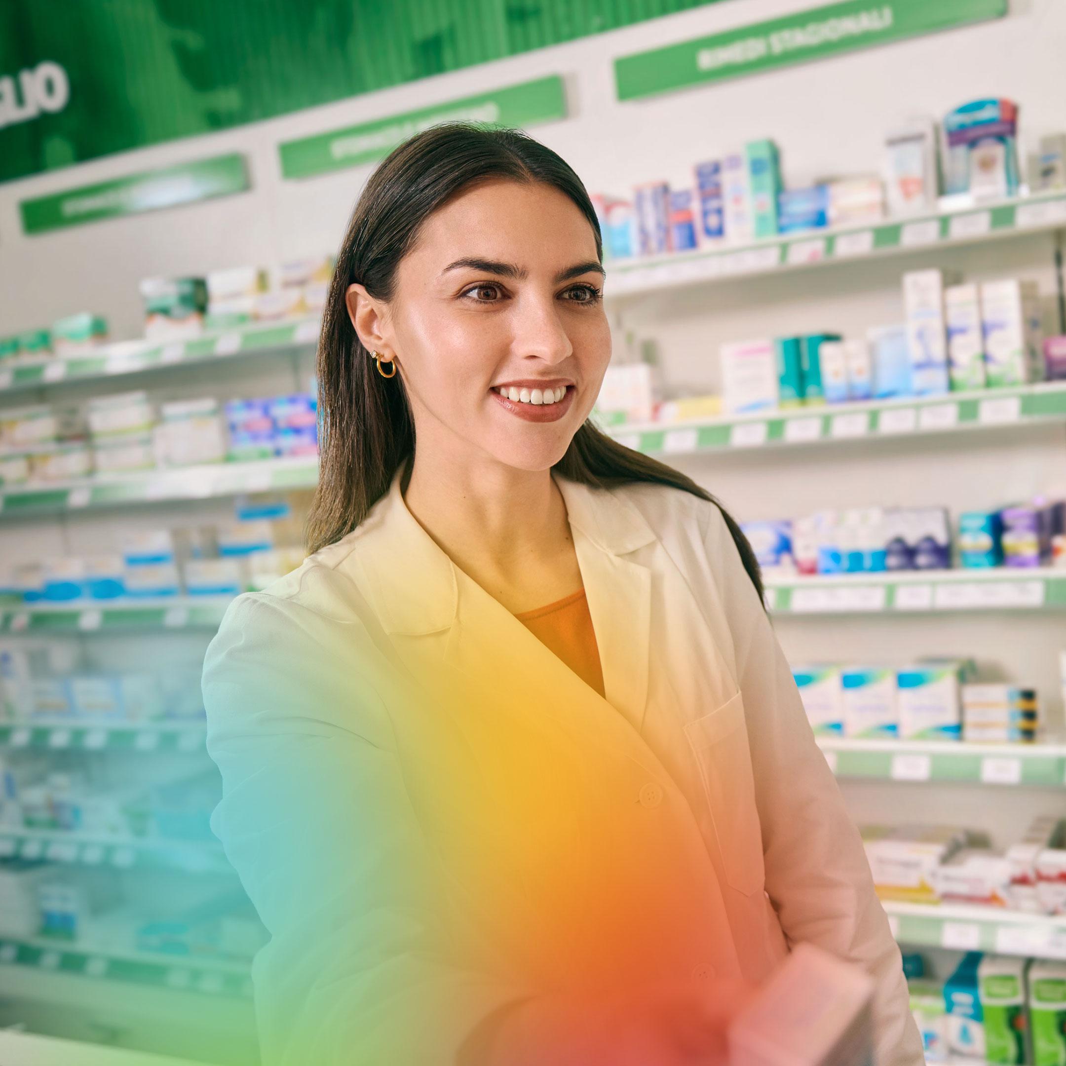 A pharmacist in a white coat handing a medication package to a customer in a pharmacy. The background shows shelves filled with various medical and personal care products.

