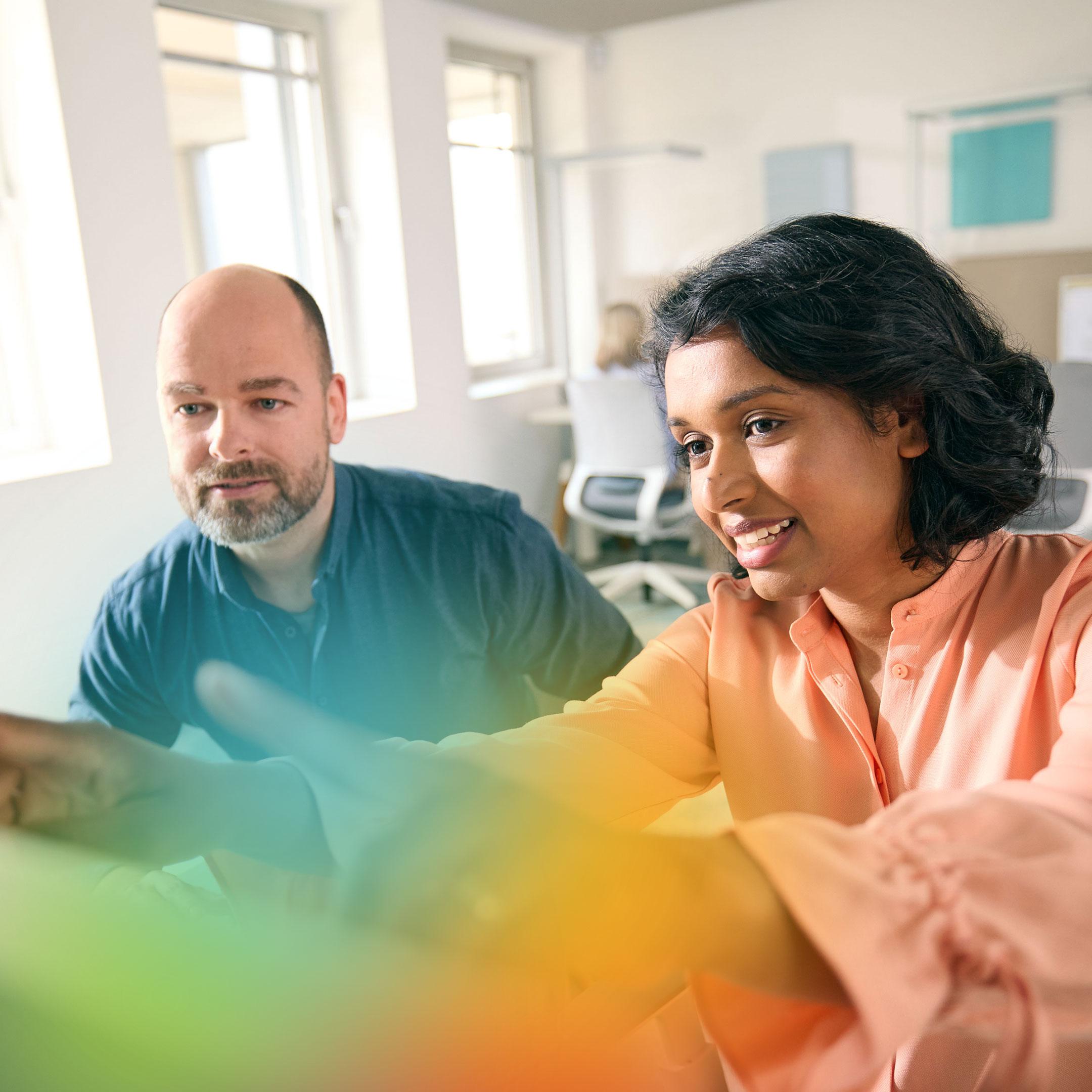 Two people collaborate in an office environment, pointing at something on a computer screen.