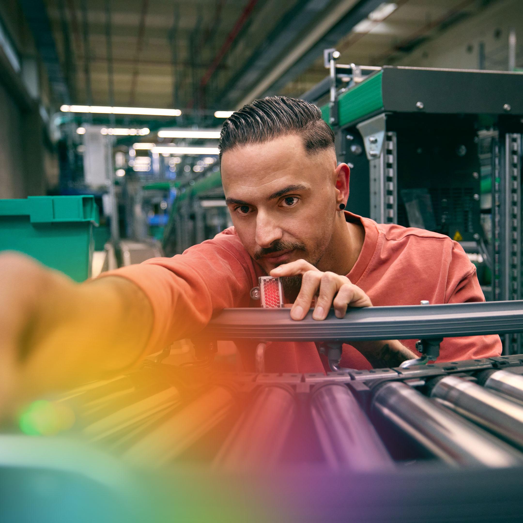 A person operating machinery in an industrial setting with various mechanical equipment and green containers in the background.