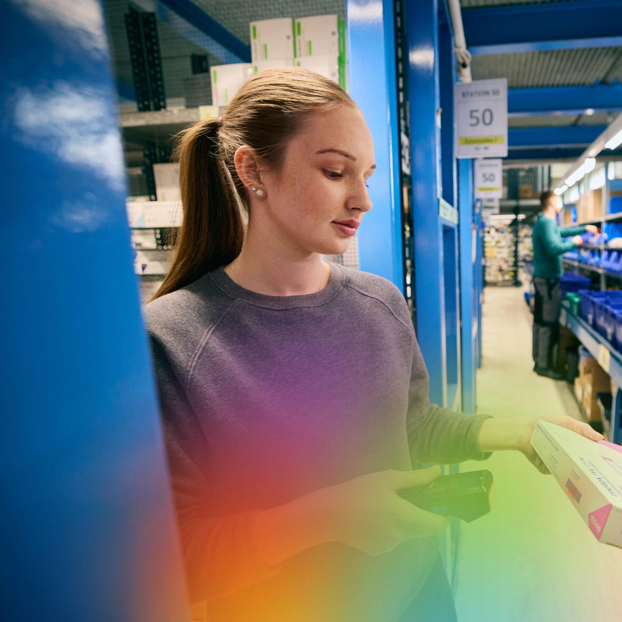 Eine Person steht in einem Lagergang und hält eine Schachtel und einen Barcode-Scanner. Im Hintergrund sind Regale mit verschiedenen Artikeln